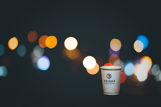 A paper cup with a logo and text sits in the foreground against a dark background filled with colorful bokeh lights. The lights are out of focus, creating a soft, luminous effect in various colors, including orange, blue, and white.