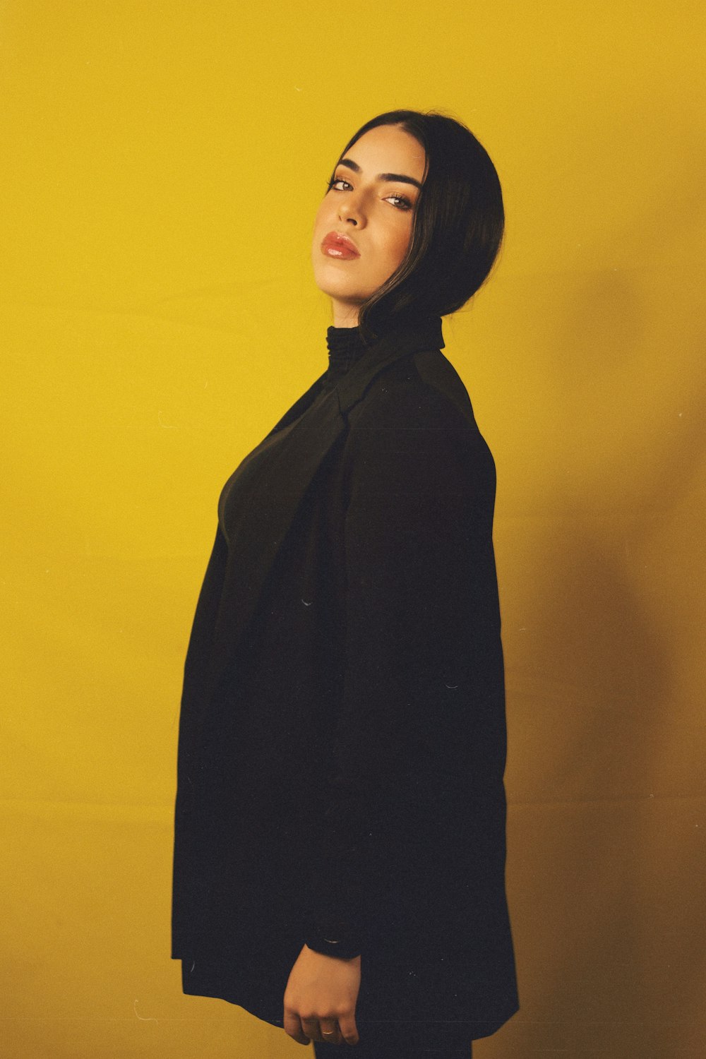 woman in black coat standing beside yellow wall