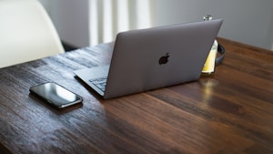 A sleek laptop with a glowing logo sits on a wooden table beside a smartphone and a partially visible juice bottle. The setting is minimalist and composed with a focus on technology and convenience.
