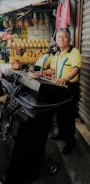 A man stands at a keyboard setup with a microphone in front of a market stall displaying various fruits such as pineapples and melons. The background includes a woman sitting and colorful clothing hanging. The man appears to be performing or practicing music.