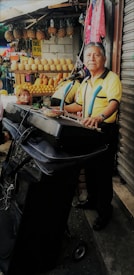 A man stands at a keyboard setup with a microphone in front of a market stall displaying various fruits such as pineapples and melons. The background includes a woman sitting and colorful clothing hanging. The man appears to be performing or practicing music.