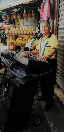 A man stands at a keyboard setup with a microphone in front of a market stall displaying various fruits such as pineapples and melons. The background includes a woman sitting and colorful clothing hanging. The man appears to be performing or practicing music.