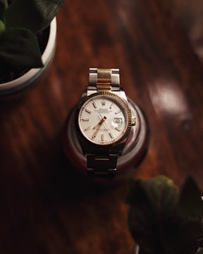A luxury wristwatch with a metal and gold band is prominently displayed on a dark wooden surface. The watch face features a classic design with Roman numerals and a date window. In the foreground, two blurred potted plants are partially visible, adding depth to the composition.