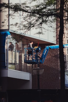 A person stands on a cherry picker, operating near the glass facade of a building. The equipment extends from a blue hydraulic lift, with the building featuring reflective windows and sections of brick walls. Overhead, branches and leaves partially obscure the view.