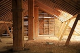 An old, rustic attic with wooden beams and walls, filled with scattered debris and straw on the floor. Light filters in through gaps in the wall slats, casting shadows and highlighting the textures of the wooden surfaces.