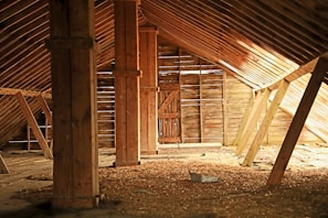 An old, rustic attic with wooden beams and walls, filled with scattered debris and straw on the floor. Light filters in through gaps in the wall slats, casting shadows and highlighting the textures of the wooden surfaces.