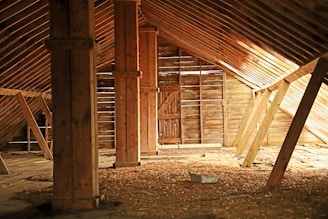 An old, rustic attic with wooden beams and walls, filled with scattered debris and straw on the floor. Light filters in through gaps in the wall slats, casting shadows and highlighting the textures of the wooden surfaces.