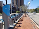 A long row of blue rental bikes is parked at a bike-sharing station along a sidewalk made of red bricks. The station is located beside a modern building with visible windows, under a clear blue sky. A tree without leaves stands along the sidewalk. Several cars are driving down the adjacent street, and a signpost with a parking sign is visible nearby.
