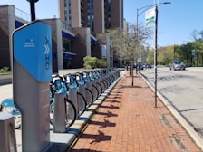 A long row of blue rental bikes is parked at a bike-sharing station along a sidewalk made of red bricks. The station is located beside a modern building with visible windows, under a clear blue sky. A tree without leaves stands along the sidewalk. Several cars are driving down the adjacent street, and a signpost with a parking sign is visible nearby.