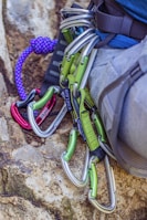 Several climbing carabiners in silver and green are attached to a harness on a person's waist, with a purple climbing rope lying nearby on a rock surface.