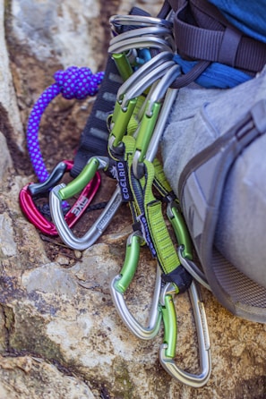 Several climbing carabiners in silver and green are attached to a harness on a person's waist, with a purple climbing rope lying nearby on a rock surface.