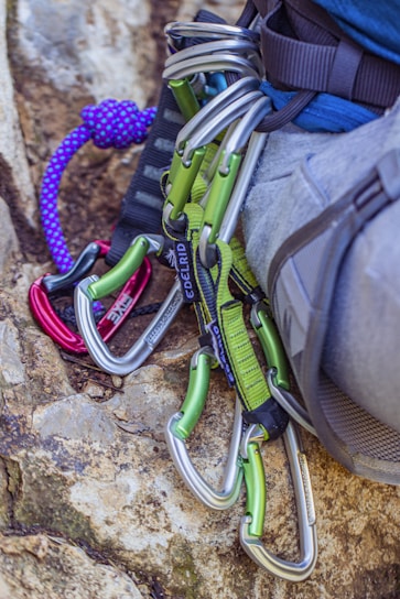 Several climbing carabiners in silver and green are attached to a harness on a person's waist, with a purple climbing rope lying nearby on a rock surface.