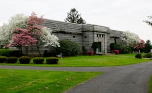 A stone mausoleum set in a manicured landscape with vibrant pink and white flowering trees on either side. The path leading up to the building is neatly paved, flanked by trimmed hedges and lush green grass. The building exudes an air of solemnity and tranquility, surrounded by natural beauty.