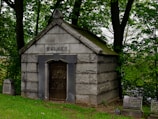 A stone mausoleum set amidst a wooded area, with the name 'Welker' engraved above its entrance. The structure features a peaked roof and a barred, arched door. Surrounding the mausoleum are several gravestones, partially covered by grass and fallen leaves. Mature trees and lush greenery add to the serene and solemn atmosphere of the setting.