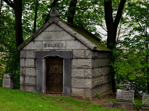 A stone mausoleum set amidst a wooded area, with the name 'Welker' engraved above its entrance. The structure features a peaked roof and a barred, arched door. Surrounding the mausoleum are several gravestones, partially covered by grass and fallen leaves. Mature trees and lush greenery add to the serene and solemn atmosphere of the setting.