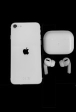 A white smartphone with an Apple logo on the back is positioned next to a pair of wireless earbuds and a charging case against a solid black background. The earbuds and case are white, matching the color scheme of the phone.
