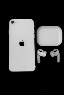 A white smartphone with an Apple logo on the back is positioned next to a pair of wireless earbuds and a charging case against a solid black background. The earbuds and case are white, matching the color scheme of the phone.