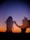Silhouetted figures dance joyfully against a vivid sunset, with the sky transitioning from deep orange to dark blue. The motion conveys a sense of freedom and spontaneity.