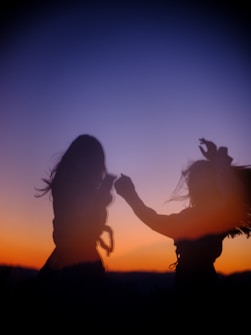 Silhouetted figures dance joyfully against a vivid sunset, with the sky transitioning from deep orange to dark blue. The motion conveys a sense of freedom and spontaneity.
