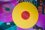 Two vinyl records are displayed, one in vibrant yellow with a red center and the other in vivid pink. The records rest atop a collection of album covers featuring various artistic designs and text, including the words 'Enter the Wu-Tang'.
