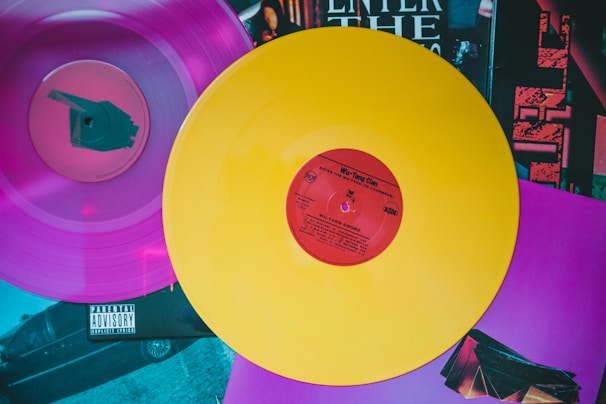 Two vinyl records are displayed, one in vibrant yellow with a red center and the other in vivid pink. The records rest atop a collection of album covers featuring various artistic designs and text, including the words 'Enter the Wu-Tang'.