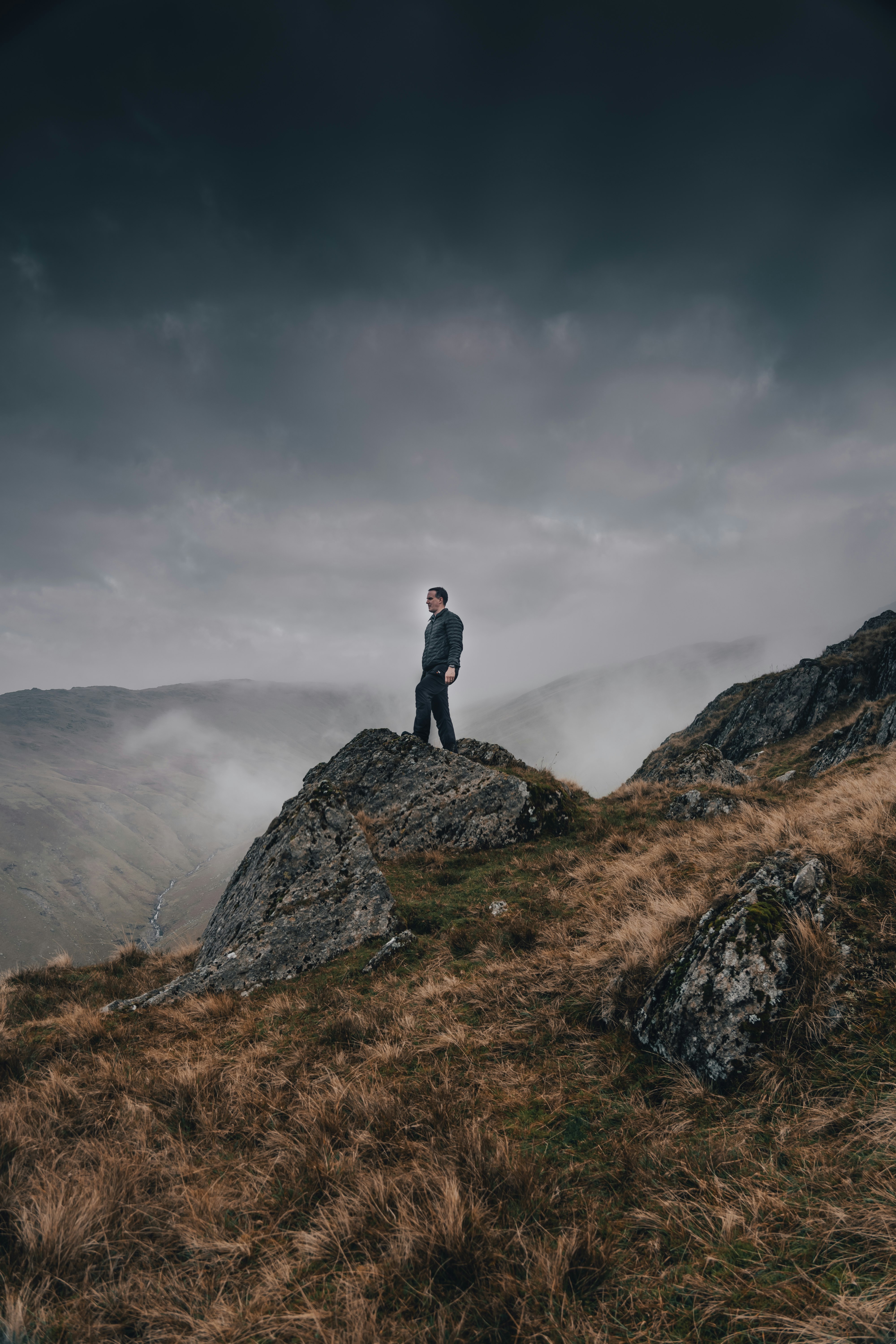 man in black jacket standing on rock mountain under gray clouds during daytime