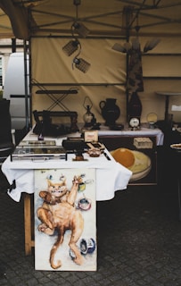 An outdoor stall displays a variety of antique and vintage items including pottery, lamps, clocks, and artworks. A prominent painting of a whimsical cat stretches its arms upward, surrounded by smaller elements painted in vibrant colors. The stall is set up under a canopy, and items are neatly arranged on tables covered with white cloth.