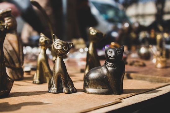 A collection of small metallic cat statues placed on a wooden surface. The statues are of different designs and finishes, with some appearing shiny and others matte. The background is blurred, indicating they are displayed outdoors, possibly at a market or fair.