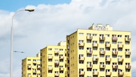 A series of yellow apartment buildings with multiple balconies and windows stand under a cloudy blue sky. A streetlamp is visible on the left side, and some graffiti is present on top of one of the buildings.