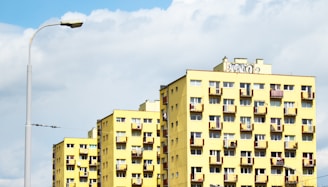 A series of yellow apartment buildings with multiple balconies and windows stand under a cloudy blue sky. A streetlamp is visible on the left side, and some graffiti is present on top of one of the buildings.