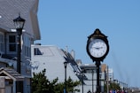 A black clock with a white face and black hands displays the time in front of buildings with gray shingled roofs. Streetlights line the sidewalk, and there are bushes and people walking in the distance under a clear blue sky.