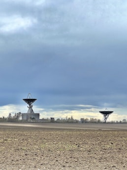 Two large satellite dishes are situated in an open, barren field with a few scattered trees in the distance. The sky above is cloudy, with a mix of grey and blue hues.
