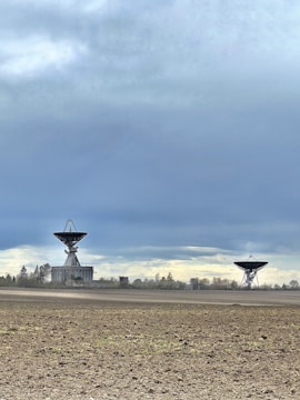 Two large satellite dishes are situated in an open, barren field with a few scattered trees in the distance. The sky above is cloudy, with a mix of grey and blue hues.