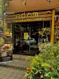 The entrance of a cozy cafe and patisserie named 'edam' with wooden signs and glass doors. Lush greenery and decorative plants frame the doorway, creating a welcoming and inviting atmosphere. A standee displays menu items like coffee and snacks, suggesting a focus on artisanal and gourmet offerings. The overall setting appears elegant and warm.