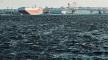 A large cargo ship with a red and white hull is navigating through dark, choppy waters, approaching a long bridge. The skyline features industrial structures and cranes in the background under a clear sky.