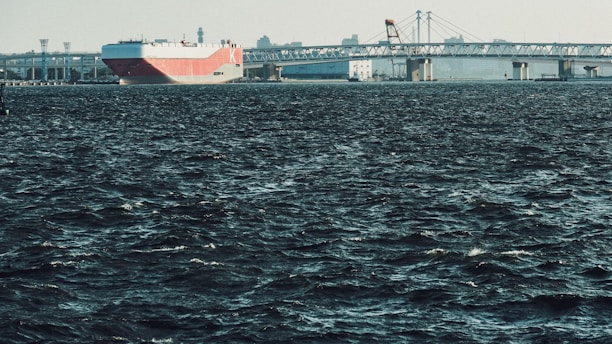 A large cargo ship with a red and white hull is navigating through dark, choppy waters, approaching a long bridge. The skyline features industrial structures and cranes in the background under a clear sky.