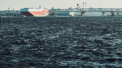 A large cargo ship with a red and white hull is navigating through dark, choppy waters, approaching a long bridge. The skyline features industrial structures and cranes in the background under a clear sky.