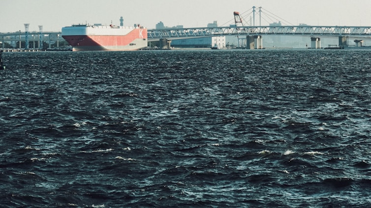 A large cargo ship with a red and white hull is navigating through dark, choppy waters, approaching a long bridge. The skyline features industrial structures and cranes in the background under a clear sky.