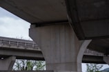 A close-up view of large concrete pillars supporting an elevated highway or bridge structure. The scene captures the geometric patterns and the texture of raw concrete. Sparse trees can be seen in the background, adding a touch of greenery to the predominantly industrial setting.
