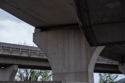 A close-up view of large concrete pillars supporting an elevated highway or bridge structure. The scene captures the geometric patterns and the texture of raw concrete. Sparse trees can be seen in the background, adding a touch of greenery to the predominantly industrial setting.