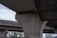 A close-up view of large concrete pillars supporting an elevated highway or bridge structure. The scene captures the geometric patterns and the texture of raw concrete. Sparse trees can be seen in the background, adding a touch of greenery to the predominantly industrial setting.