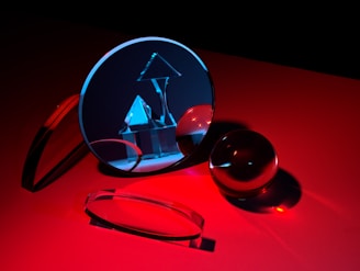 A collection of glass objects is arranged on a red surface, including a circular mirror reflecting geometric shapes such as a pyramid and cube under blue light. The scene captures reflections and refractions, resulting in a play of light and shadows.