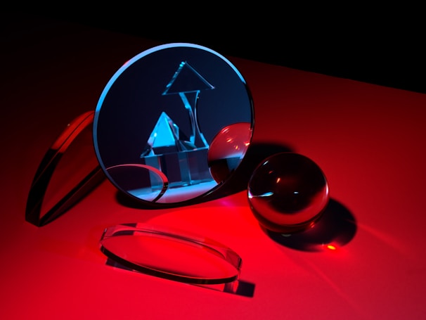 A collection of glass objects is arranged on a red surface, including a circular mirror reflecting geometric shapes such as a pyramid and cube under blue light. The scene captures reflections and refractions, resulting in a play of light and shadows.
