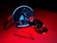 A collection of glass objects is arranged on a red surface, including a circular mirror reflecting geometric shapes such as a pyramid and cube under blue light. The scene captures reflections and refractions, resulting in a play of light and shadows.