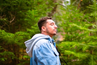 A man in a blue denim jacket with a gray hood stands with his eyes closed among lush green trees, creating a sense of serenity and connection with nature.