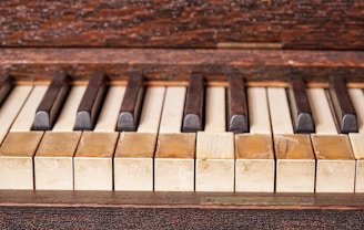 A close-up image of an old piano keyboard with worn and discolored keys, showing signs of age and use. The white keys have yellowed over time, and the black keys are slightly uneven, adding to the vintage appearance.