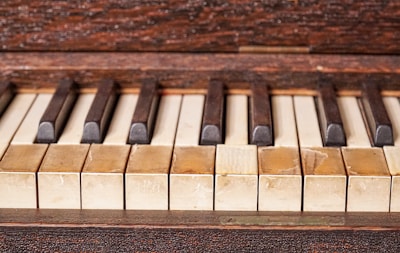 A close-up image of an old piano keyboard with worn and discolored keys, showing signs of age and use. The white keys have yellowed over time, and the black keys are slightly uneven, adding to the vintage appearance.