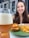 A glass of beer with a foamy head is held in the foreground. A plate with a cheeseburger and fries is placed in front of a smiling person in the background.
