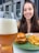 A glass of beer with a foamy head is held in the foreground. A plate with a cheeseburger and fries is placed in front of a smiling person in the background.