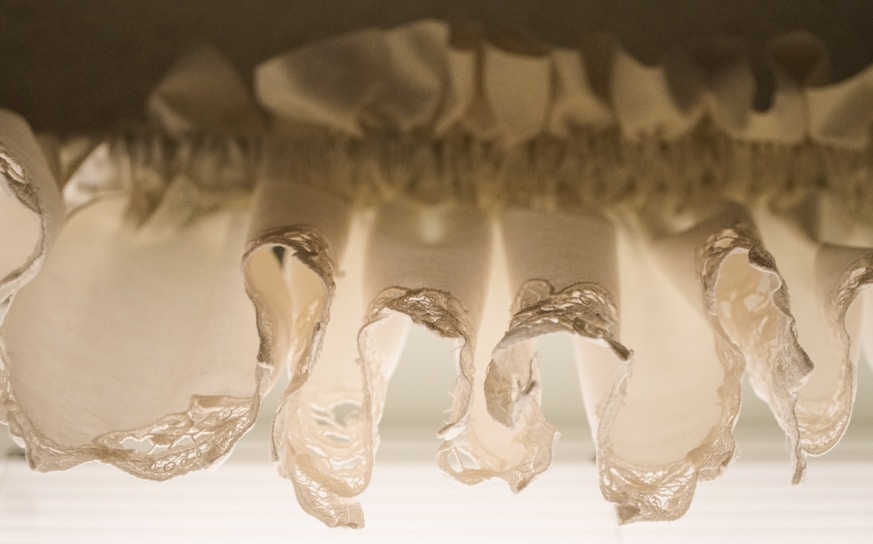 Close-up view of delicate, beige fabric ruffles with ornate lace edges, gathered in a flowing, wavy pattern.